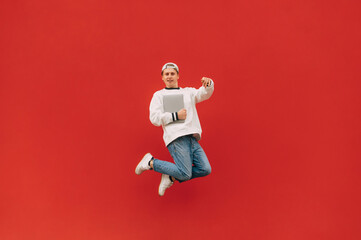 Cheerful guy in stylish casual clothes jumps with a laptop in his hands on a red background and looks at the camera with a smile on his face.