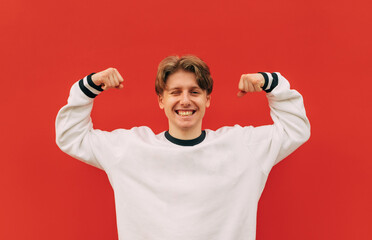 Joyful young man in light casual clothes stands on a background of red wall and shows a joking biceps and looks at the camera with a smile on his face.