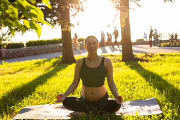 Cute young caucasian pregnant woman is meditating while sitting on a rug on the lawn on a sunny...