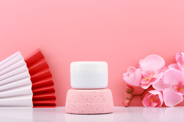 Fototapeta na wymiar White cosmetic jar on pink pedestal against pink background with copy space decorated with wavers and flowers. Concept of beauty product, hair mask, cream or scrub for skin care.