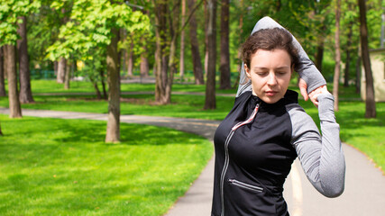 Brunette sporty girl stretching arms before training, workout in green city park, wearing sportwear. Exercising outdoors before jogging. Preparing muscles and ligaments. Copy space.