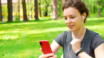 Pretty young woman looking at smartphone, sitting on green grass in city park outdoors, using headphones. Female student watching online webinar, video, listening audio course, podcast ,social media
