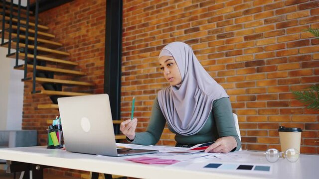 Muslim girl designer has a video call from her office. A woman holds a pencil and samples of materials in her hands. Progressive woman in hijab