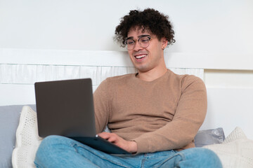Happy positive guy, young cheerful joyful man is relaxing, enjoying, lying on bed, using his laptop...