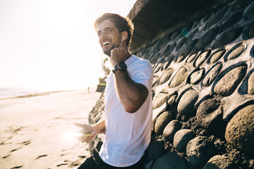Cheerful man standing with headphones and smartphone on sandy coast