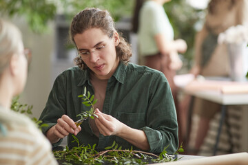 Portrait of young male florist cutting green plants while creating floral compositions for decor,...
