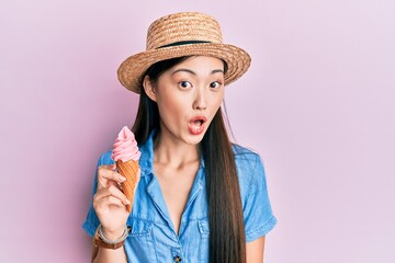 Young chinese woman wearing summer hat holding ice cream scared and amazed with open mouth for surprise, disbelief face