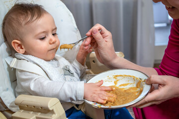 Charming messy little baby boy sitting in a feeding chair, eating and playing with his first food from the spoon with mom at home. 
