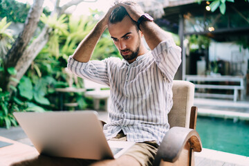 Young serious bearded businessman touching head and doing remote work