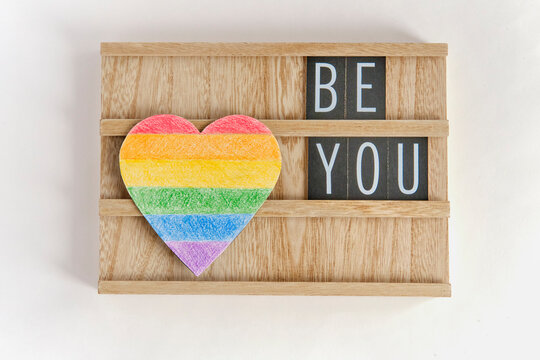 Paper heart painted with colored pencils like a LGBT flag and text Be you on a wooden stand on white background.