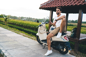 Cheerful masculine traveler with smartphone resting on scooter in countryside