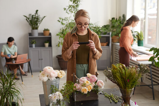 Portrait of young female florist taking photos of floral compositions via smartphone while working in flower shop, copy space
