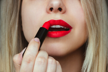 Cropped shot of a young beautiful caucasian blonde painting her lips with red lipstick on a dark background. Close-up - obrazy, fototapety, plakaty