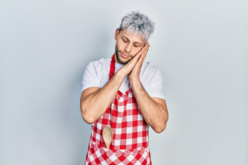 Young hispanic man with modern dyed hair wearing apron sleeping tired dreaming and posing with hands together while smiling with closed eyes.