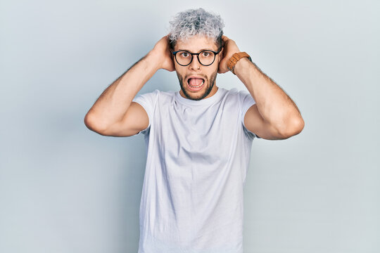 Young hispanic man with modern dyed hair wearing white t shirt and glasses crazy and scared with hands on head, afraid and surprised of shock with open mouth