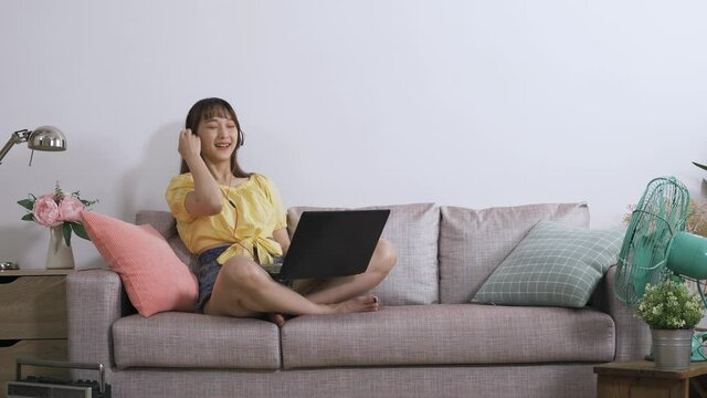 Asian Girl Wearing Headphones Is Leaning Backward And Covering Her Mouth While Watching A Funny Video On Computer In A Modern Living Room With White Wall.