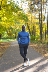Fototapeta premium Cheerful african american young man in stylish clothes walks in autumn park on sunny warm autumn day. Concept of weekend and outdoor recreation