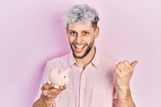 Young hispanic man with modern dyed hair holding piggy bank pointing thumb up to the side smiling happy with open mouth