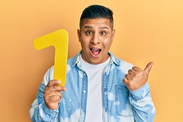 Young latin man holding number one pointing thumb up to the side smiling happy with open mouth