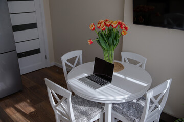 A bouquet of bright tulips on the table. Laptop and tulips in the interior of a bright kitchen. Spring flowers. Fresh orange buds. Work from home. Cozy kitchen