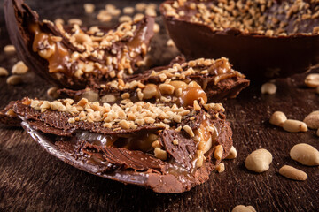 Stuffed chocolate easter egg with peanut and caramel on a wooden table with lots of peanuts around. Cracked. Close up.