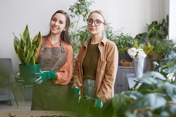 Obraz premium Waist up portrait of two young female florists smiling at camera while potting plants in flower shop or gardening together, copy space