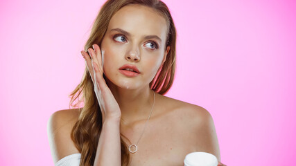 young woman looking away while applying face cream on pink