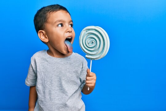 Adorable latin toddler eating delicious lollipop over isolated blue background.