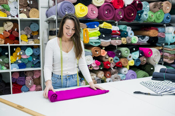 Young brunette woman seller standing near work place in fabric store. Owner textile shop packing tissue on table. Small business concept