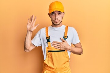 Hispanic young man wearing handyman uniform swearing with hand on chest and open palm, making a...