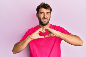 Handsome man with beard making heart symbol with hands shape clueless and confused expression....
