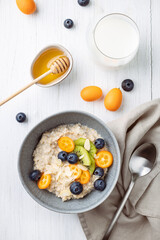 Oatmeal porridge with kumquat, blueberries, kiwi and almond flakes in a bowl. Healthy breakfast with milk and honey on a white table.