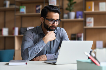 Young serious indian professional business man, focused ethnic male student wearing glasses working on laptop, remote studying using computer looking at screen watching seminar webinar at home office.