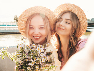 Two young beautiful smiling hipster woman in trendy summer sundress. Sexy carefree women posing on the street background in hats. Positive models at embankment at sunset. Taking selfie.Holding flowers