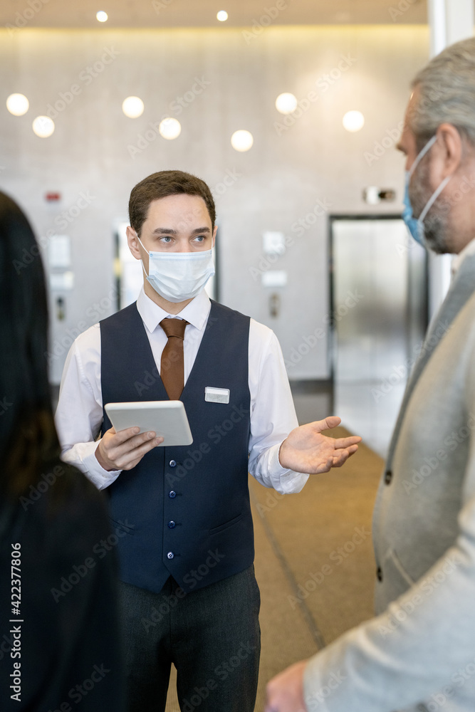 Aufkleber Young male administrator of hotel in protective mask and formalwear using touchpad while talking to new guests in the lounge