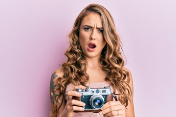 Young blonde girl holding vintage camera in shock face, looking skeptical and sarcastic, surprised with open mouth