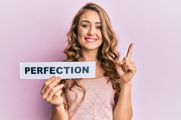Young blonde girl holding paper with perfection message smiling with an idea or question pointing...