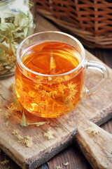 Linden herbal tea with flowers in a glass cup on wooden rustic board with lime blossom dry herb nearby, closeup, drink for relaxation, mental health,  naturopathy, homeopathy and folk medicine concept