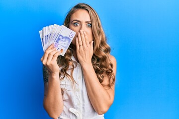 Young blonde girl holding swedish krona banknotes covering mouth with hand, shocked and afraid for mistake. surprised expression