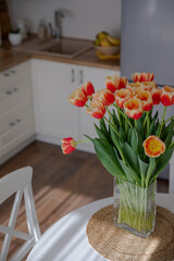 A bouquet of bright tulips on the table. A bright doll in the interior of a light kitchen. Spring flowers. Fresh orange buds. Cozy kitchen