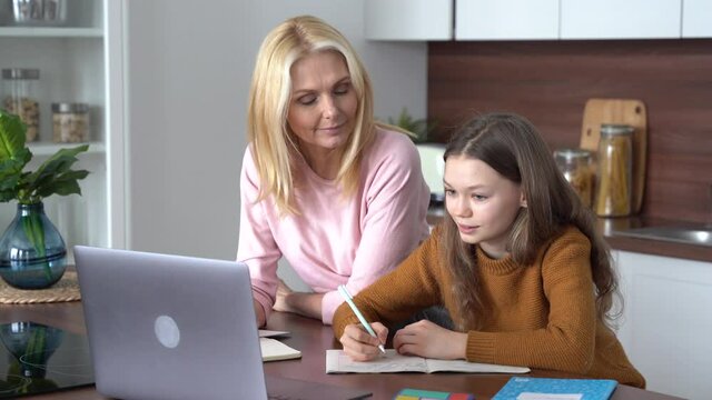 Teen child daughter studying at home in kitchen with mom. Teenage school kid girl distance learning virtual online class with mother or tutor helping doing homework together during remote education.