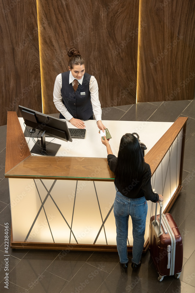 Poster young receptionist in uniform standing by workplace and passing passport to female traveler over rec