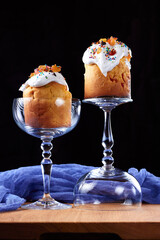 Two Easter cakes (sweet bread Orthodox kulich, paska) decorated with white frosting and candy sprinkles on glass beakers on a dark background