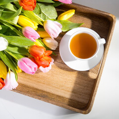 A bouquet of multicolored tulips and a cup of tea are on a tray on the table