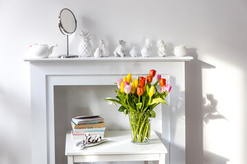 Bouquet of colorful tulips in a glass figured vase on a white fireplace console. A faience figurine of angel as an interior decoration.
