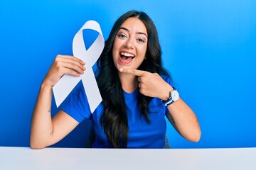 Beautiful brunette young woman holding white ribbon smiling happy pointing with hand and finger