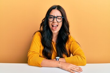 Beautiful brunette young woman wearing glasses and casual clothes sitting on the table winking...