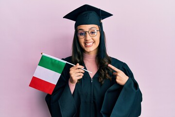 Young hispanic woman wearing graduation uniform holding italy flag smiling happy pointing with hand and finger