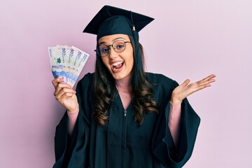 Young hispanic woman wearing graduation uniform holding colombia pesos banknotes celebrating achievement with happy smile and winner expression with raised hand