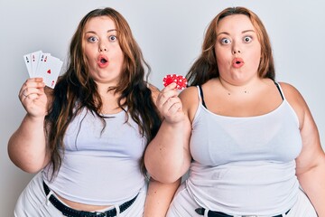 Plus size caucasian sisters woman holding poker chips and cards scared and amazed with open mouth for surprise, disbelief face